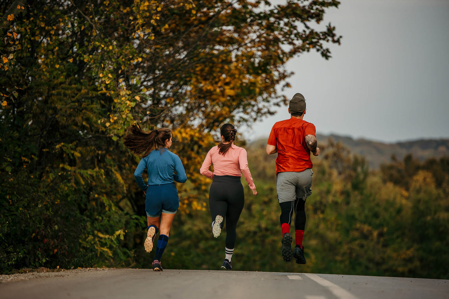 Vue de dos de trois coureurs adultes faisant un marathon sur une route de campagne bordée d’arbres aux feuilles d’automne.