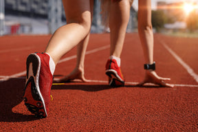 Gros plan sur une chaussure de course rouge à crampons sur une piste d’athlétisme synthétique, avec une athlète en position de départ prête à s’élancer pour un 10 km.