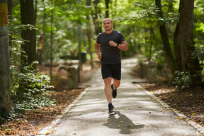 Un homme chauve en tenue de sport grise et noire courant un 5 km sur un chemin pavé entouré d’une forêt verdoyante.