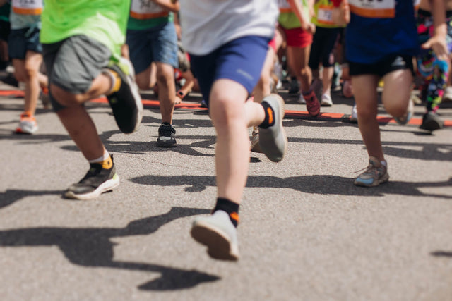 Jambes de plusieurs enfants en mouvement lors d’une course sur asphalte, portant des tenues de sport colorées et des espadrilles.